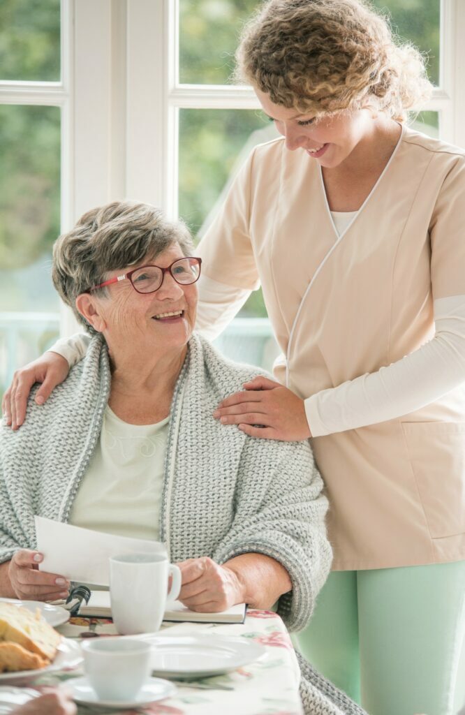 Positive senior lady siting at table and smiling to her young nurse