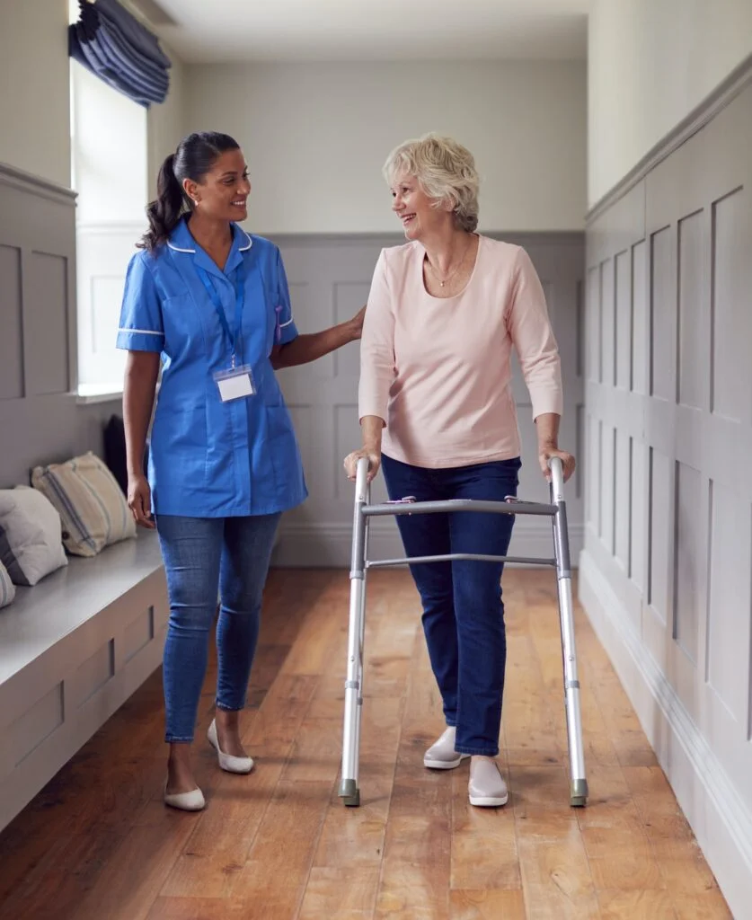 Senior Woman At Home Using Walking Frame Being Helped By Female Care Worker In Uniform